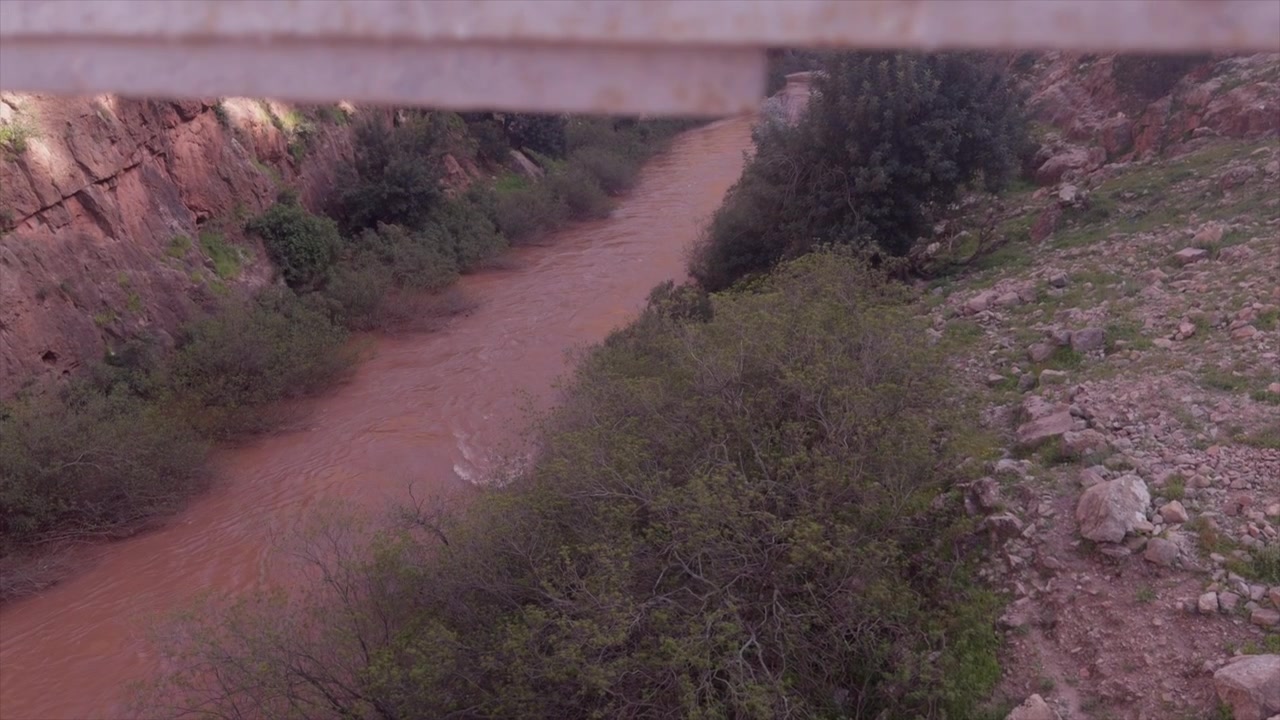 Muddy red river winds through rocky canyon walls and green shrubs viewed from bridge