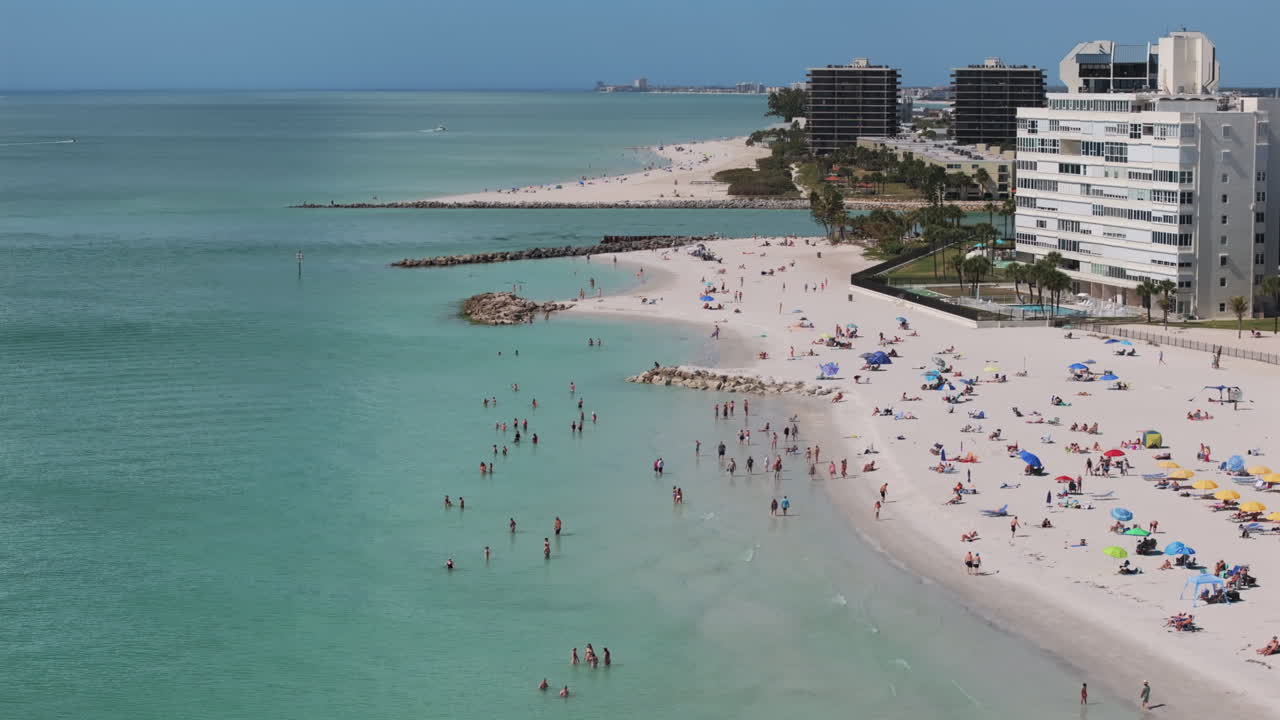 Vista aerea dell'affollata spiaggia di Upham a Saint Pete Beach che mostra bagnanti, ombrelloni colorati, nuotatori nelle acque turchesi del Golfo, un molo frangiflutti e condomini fronte mare in una popolare località turistica della Florida