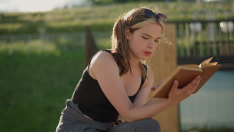 woman seated with legs crossed, reading a book while the wind blows her hair and the book s pages, a black bag is beside her, and the blurred background features a lush green hill