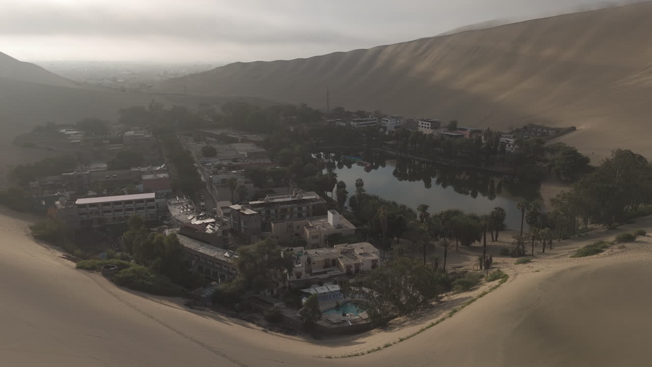 Drone shot flying over the city of Huacachina in Peru behind the dunes on a cloudy day during the mist in the morning with the sun coming through LOG