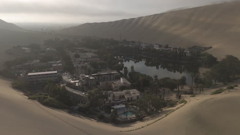 drone shot flying over the city of huacachina in peru behind the dunes on a cloudy day during the mist in the morning with the sun coming through log