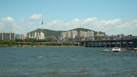 Hermoso-Día-Soleado-Cerca-Del-Río-Han,-Torre-Namsan-Y-Puente-Banpo-De-Dos-Pisos-En-El-Fondo,-Catamarán-Y-Velero-Flotando-En-El-Muelle