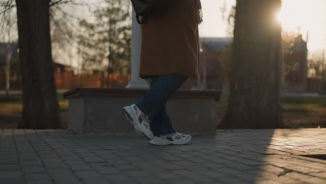 a person wearing a brown coat, jeans, white shoes, and a backpack is shown walking slowly through a park at sunset. the focus is on their legs, emphasizing the fainted steps