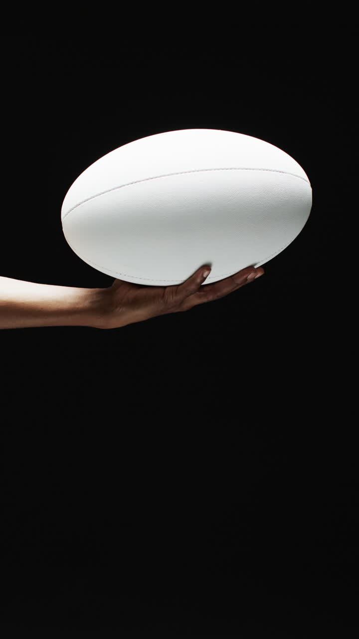 Vertical video of hand of african american man holding white rugby ball on black background