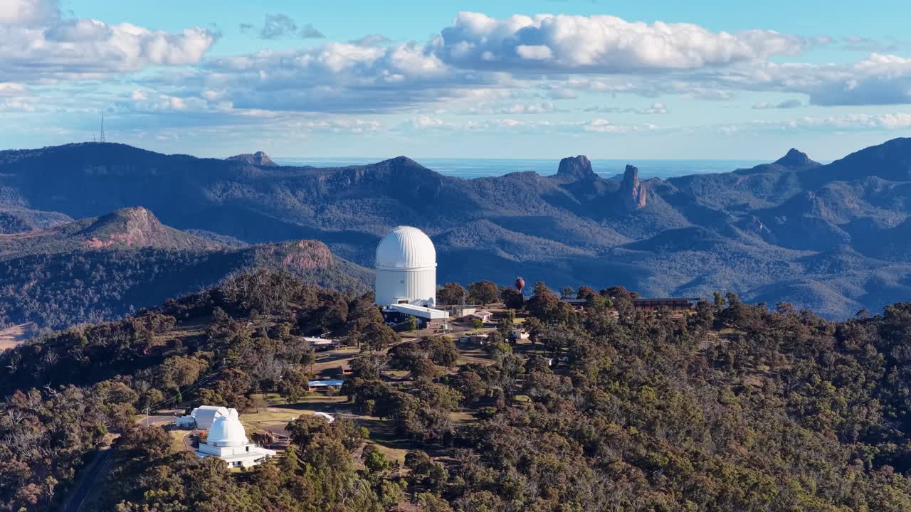 Drone camera orbits a large astronomical observatory dome atop a forested ridge, revealing rugged mountain landscape and dramatic sunset lighting in Warrumbungle National Park