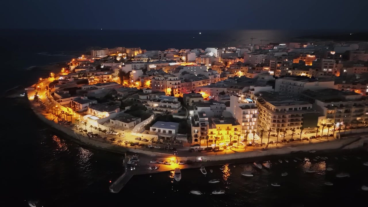 Marsaskala-bukten, Maltas strandpromenad med upplysta båtar, yachter och bostadshus i skymningen, reflektioner på Medelhavets strandlinje i en drönarvy