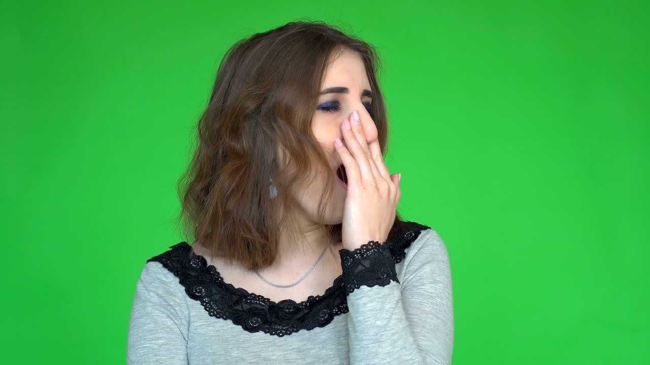 Tired young woman yawns and covering her mouth while looking at the camera over green background.