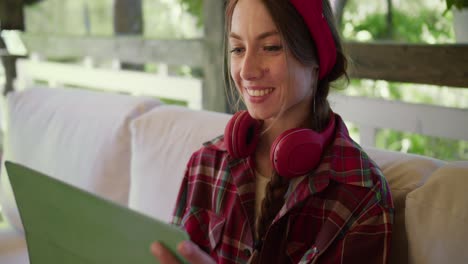 Close-up-shot:-a-girl-in-red-clothes-and-red-headphones-sits-on-a-sofa-and-works-on-a-green-tablet-in-a-gazebo-in-nature