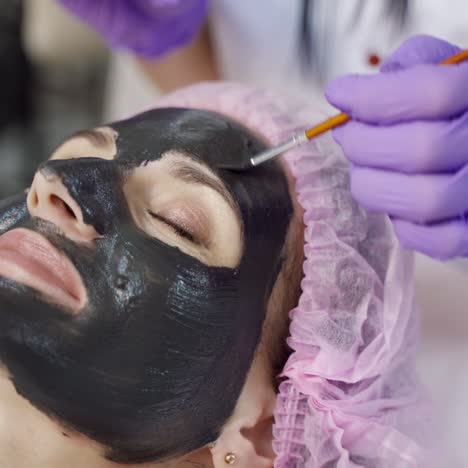 a young woman is lying on a couch with a black mask on her face, surrounded by a beautician which applying it in a beauty salon. close-up.