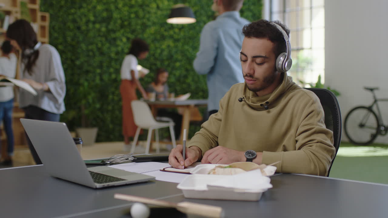 young arab businessman student brainstorming writing notes enjoying study using laptop researching online listening to music in busy office workplace