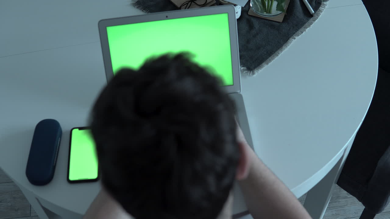 Close-up the man sits at the table and uses a computer and phone, green screen, white table with electronics, top view