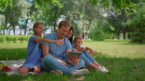 Smiling-family-waving-hands-communicate-using-tablet-on-nature-in-summer-day.