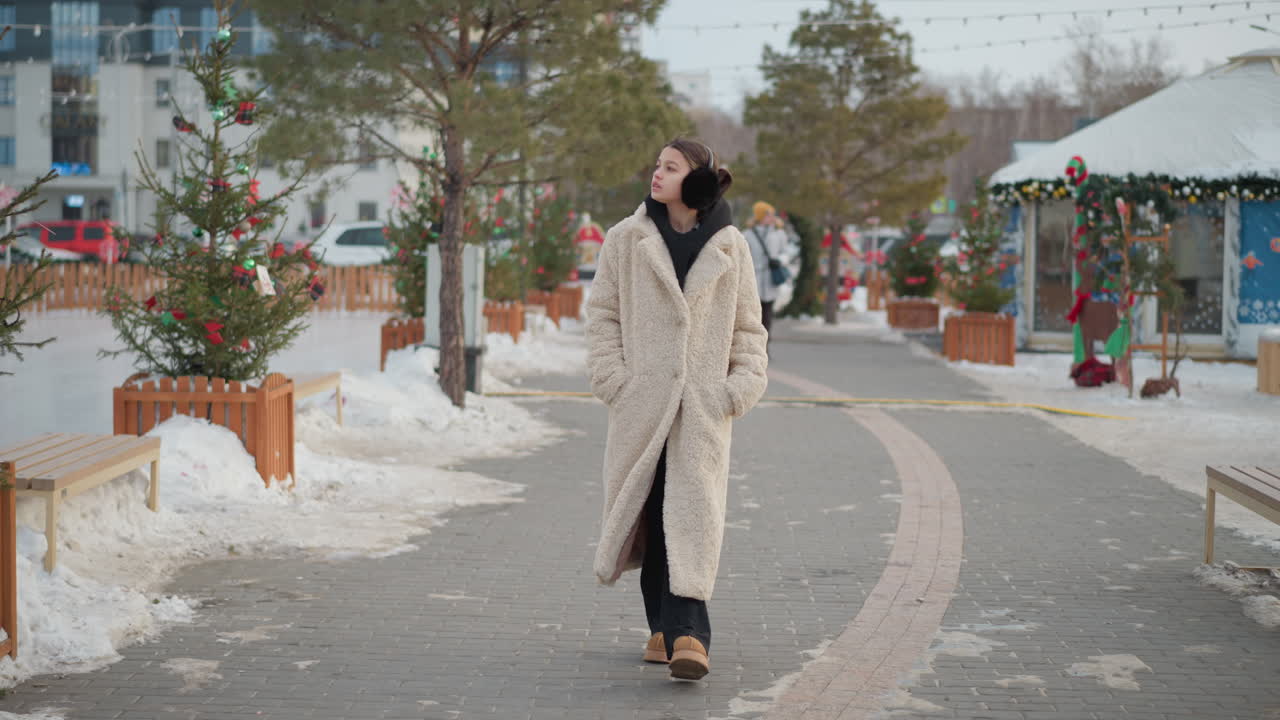 검은색 후디와 이어마프와 함께 따뜻한 베이지 겨울 코트를 입은 소녀는 주머니 안에 손을 넣고 눈 인 길을 고 있습니다. 그녀는 축제 거리를 산책하는 동안 위로 쳐다보고 있습니다.
