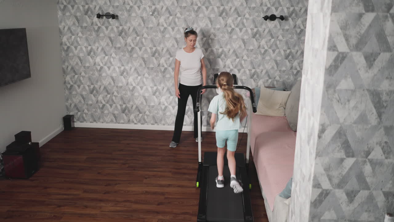 Training supervisor observes kid exercising on treadmill in modern room featuring television, patterned accent wall, and home theater system, creating calm and structured indoor fitness atmosphere