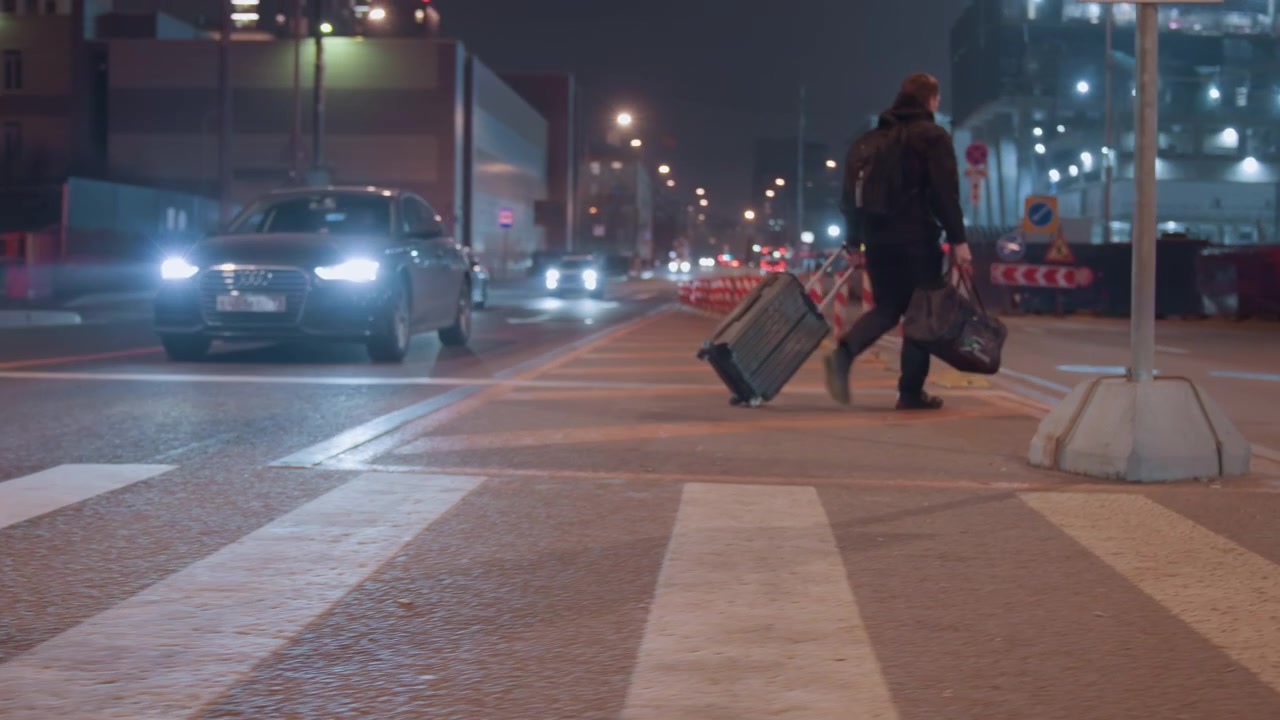 Night crosswalk with suitcase and headlights, lone traveler drags rolling luggage across striped pavement while cars wait with bright beams, construction cones and building lights frame scene