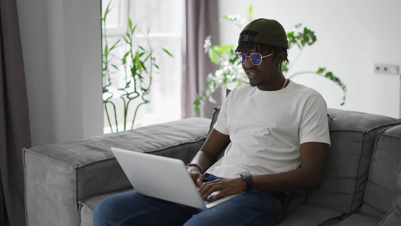 Attractive african American man in glasses sitting on the sofa using laptop at home
