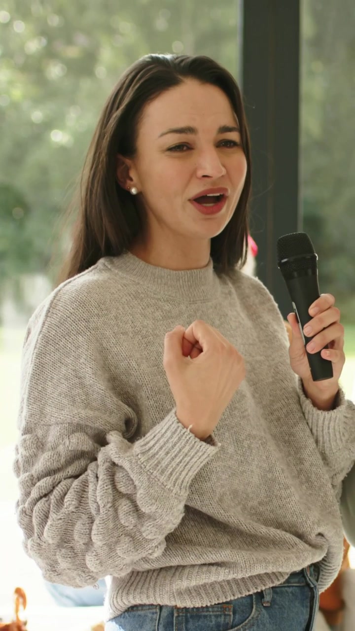 Video verticale: Donna di mezza età che canta, tenendo un microfono in maglione vicino alla finestra, enfatizzando il testo.