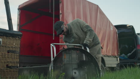 man wearing cap and gloves leans over fan grate on grassy field warming hot air balloon engine before sunrise flight as crew and vehicles stand under pastel sky over farmland