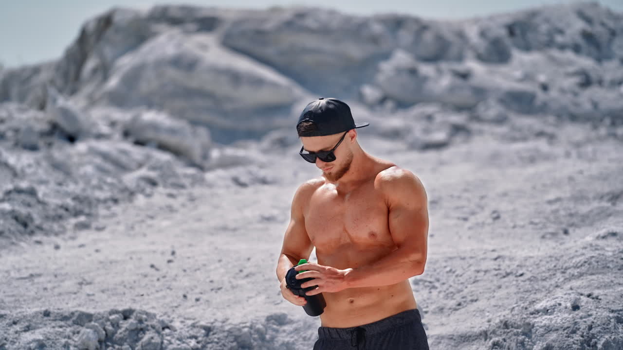 Topless sportsman drinking cold drink on natural background. Muscular man in black sunglasses drinks water after workout in the mountains.