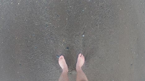 Waves-washing-up-at-pale-male-white-caucasian-legs-on-a-beach---First-person-view-looking-down-at-feet-in-sand