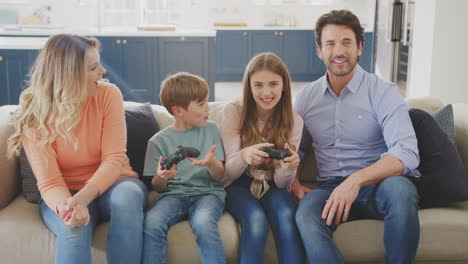 Parents-Having-Fun-Sitting-On-Sofa-With-Children-At-Home-Playing-On-Games-Console