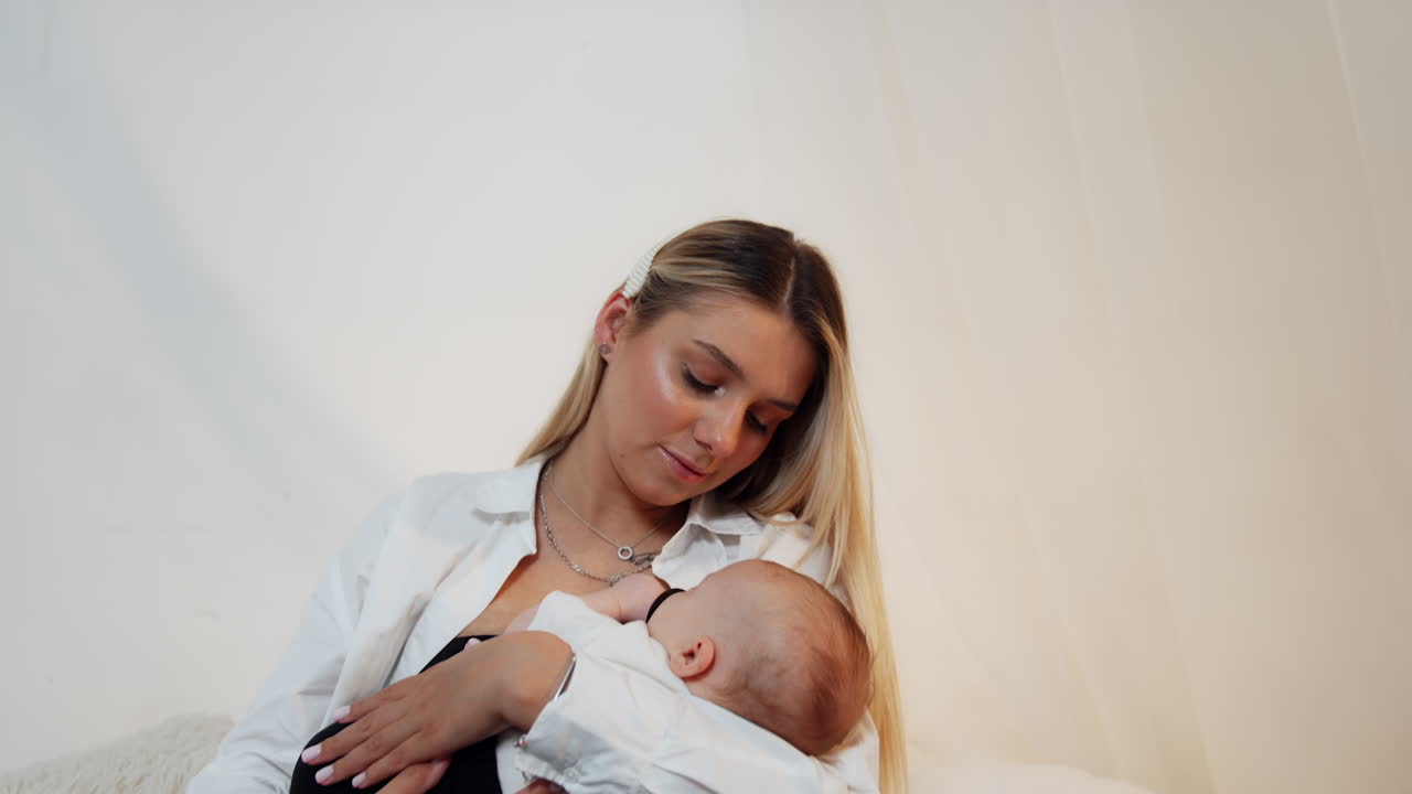 Beautiful blonde long-haired woman sits on canopy bed. Loving mom waves her adorable newborn in hands.