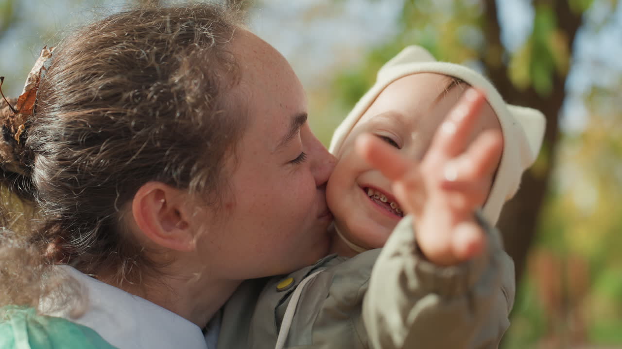 Madre caucasica bacia il bambino che allunga la mano, un bacio tenero e una mano tesa, un cappello morbido e un cappotto accogliente, uno sfondo autunnale illuminato dal sole, un legame familiare intimo e un caldo momento affettuoso nel parco.