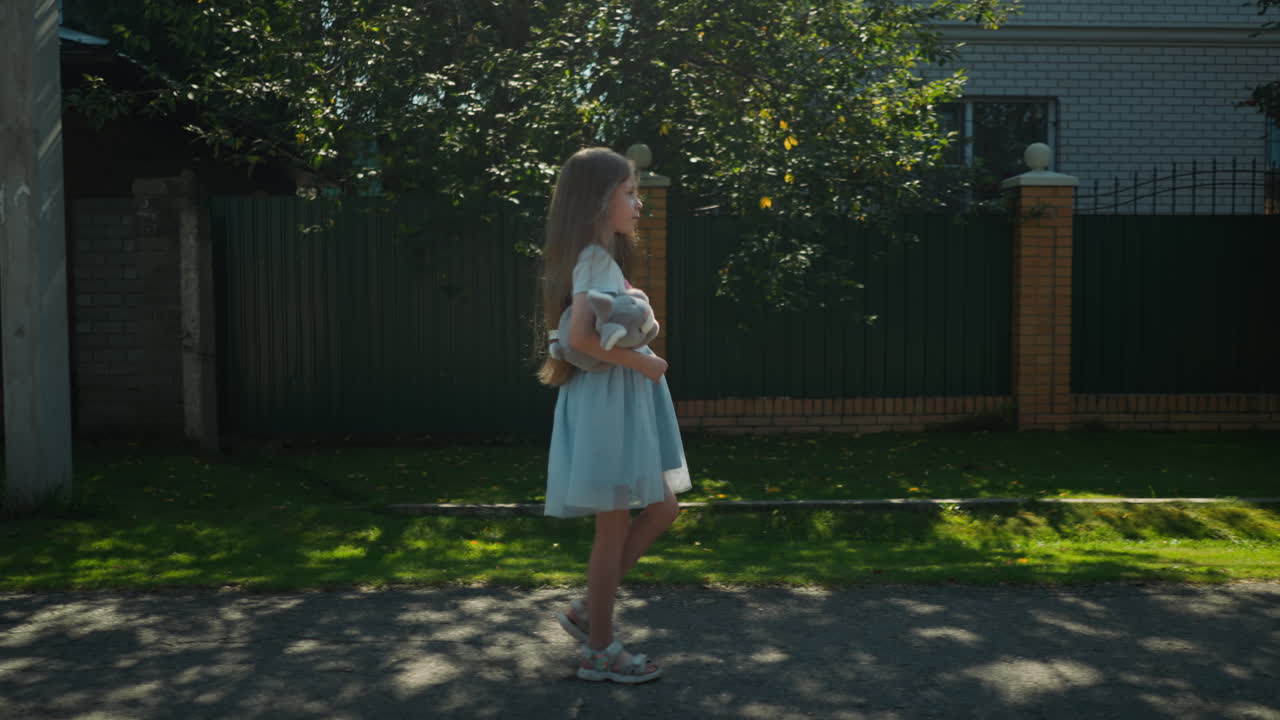 Side view of girl in blue gown with net holding teddy bear walking peacefully along quiet residential street as sun casts soft shadow on pavement and lush greenery frames background