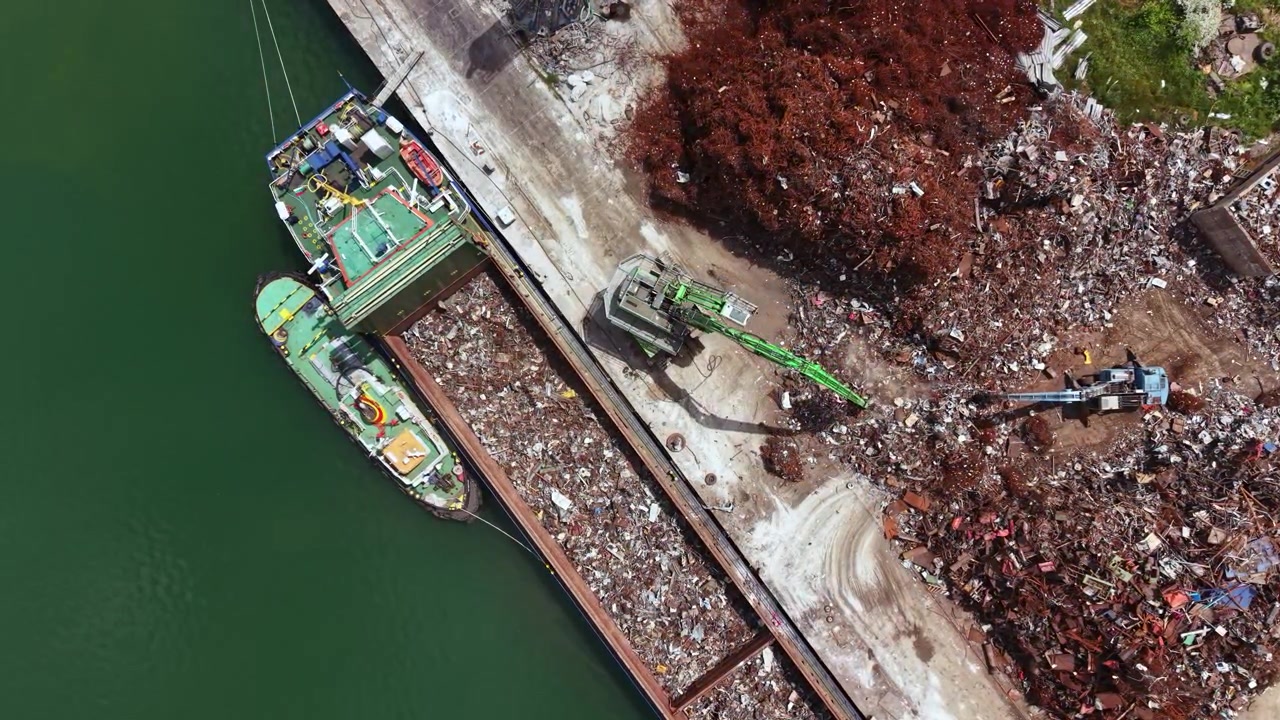 Attività di costruzione si svolge in un sito di riciclaggio vicino a un lungomare, con barche e macchinari coinvolti nella raccolta e lavorazione dei materiali durante il giorno.