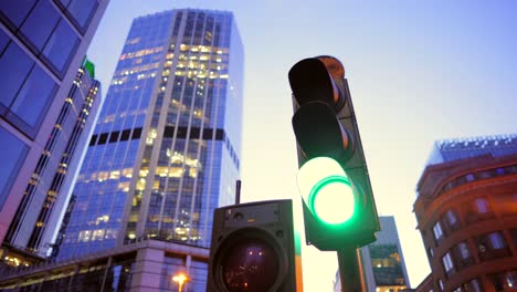 Red-traffic-light-turning-green-in-downtown-London,-at-dusk-with-building-lights,-and-double-decker-red-bus-passing-by