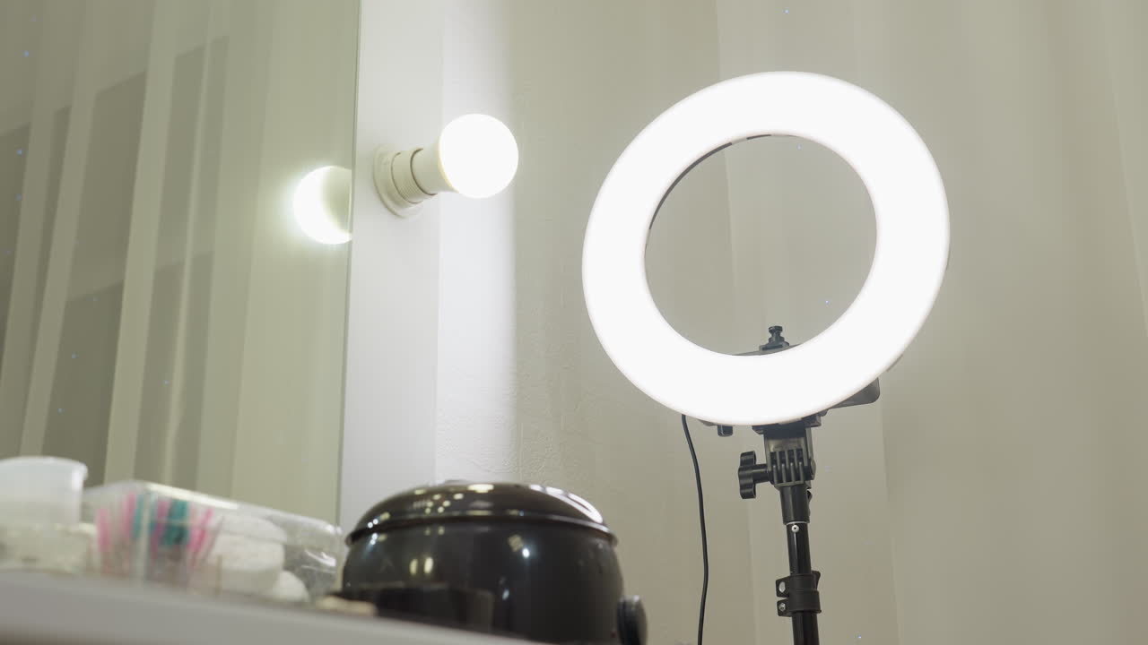 Ring light standing beside mirror and glowing bulb above organized salon workstation with beauty tools, containers, brushes, cotton pads, and product bottles neatly placed on white tabletop
