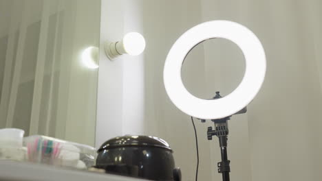 ring light standing beside mirror and glowing bulb above organized salon workstation with beauty tools, containers, brushes, cotton pads, and product bottles neatly placed on white tabletop