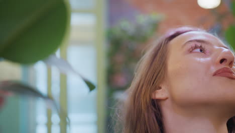 closeup of young woman with wavy hair gently reaching up to touch leaf while smiling in softly lit indoor space with blurred background featuring plants and glass doors, expressing natural joy