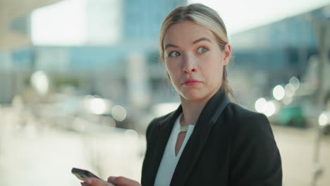 lady in professional setting operating phone stares at camera with surprised look and subtle smile, dressed in formal outfit, standing outdoors in urban environment with soft blur city background