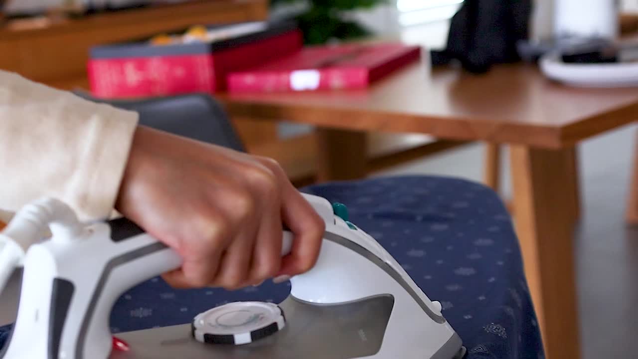 A close-up shot shows a person's hand moving a steam iron across a blue garment