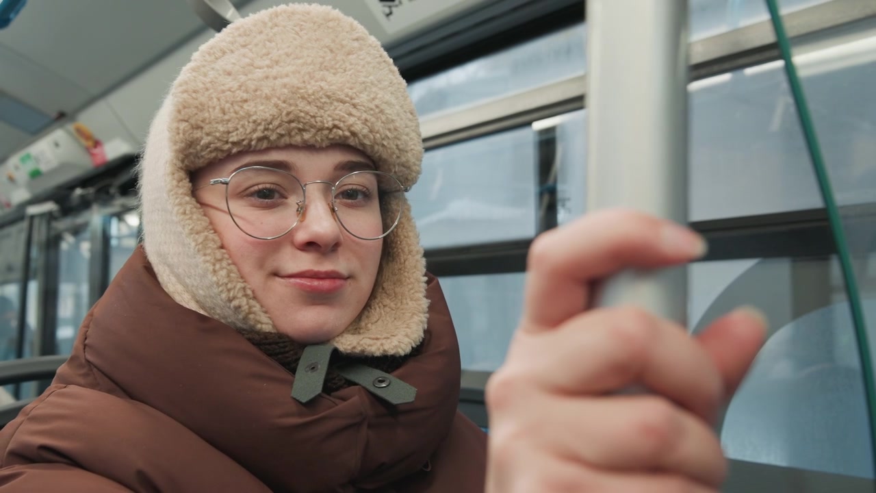 Primo piano medio di una giovane donna seduta su un autobus urbano in inverno, che afferra un palo verticale per l'equilibrio accanto alla luce della finestra, indossando un cappello da trapper in pile e un cappotto imbottito, con un sorriso calmo e gentile.