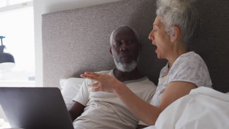 mixed race senior couple using laptop while lying in the bed at home