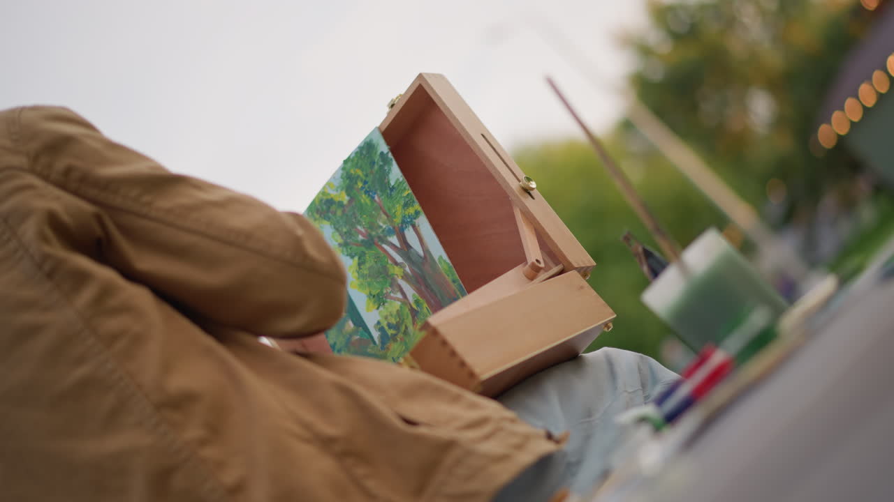 Un artista di strada bianco spacchetta un cavalletto di legno rivelando un vivace dipinto paesaggistico all'interno di una scatola portatile sul marciapiede vicino al parco, bokeh serale delicato, pennelli e palette nelle vicinanze, umore contemplativo, urbano