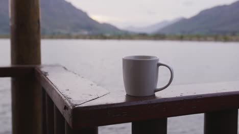 aussicht auf einen see und berge bei sonnenuntergang an einem bewölkten tag von einem log-cabin-fenster aus mit einem becher