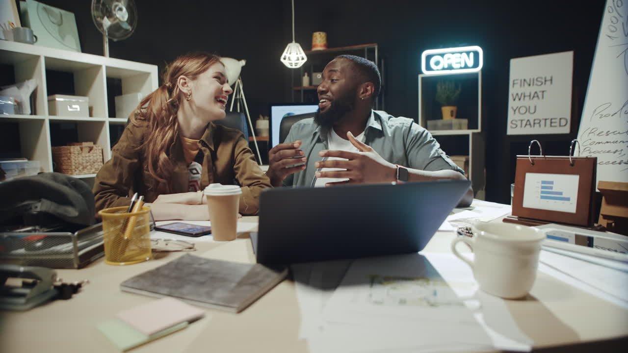 Joyful afro man and woman laughing in front of laptop screen at workplace.