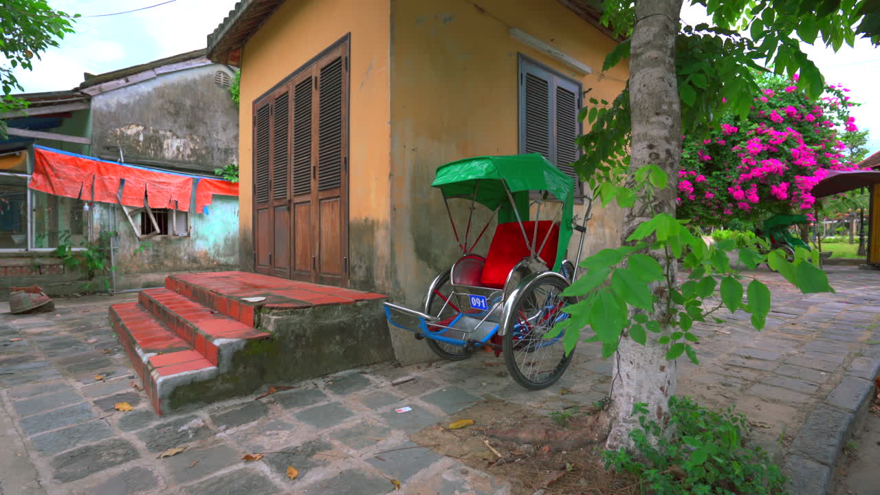 Empty rickshaw waits near vibrant building and tropical plants in Hoi An, Vietnam