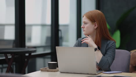 A-young-red-haired-business-woman-looks-thoughtfully-at-the-screen-and-brainstorms.-Watch-and-think-about-problems-looking-out-the-window.-Thoughtful-business-woman