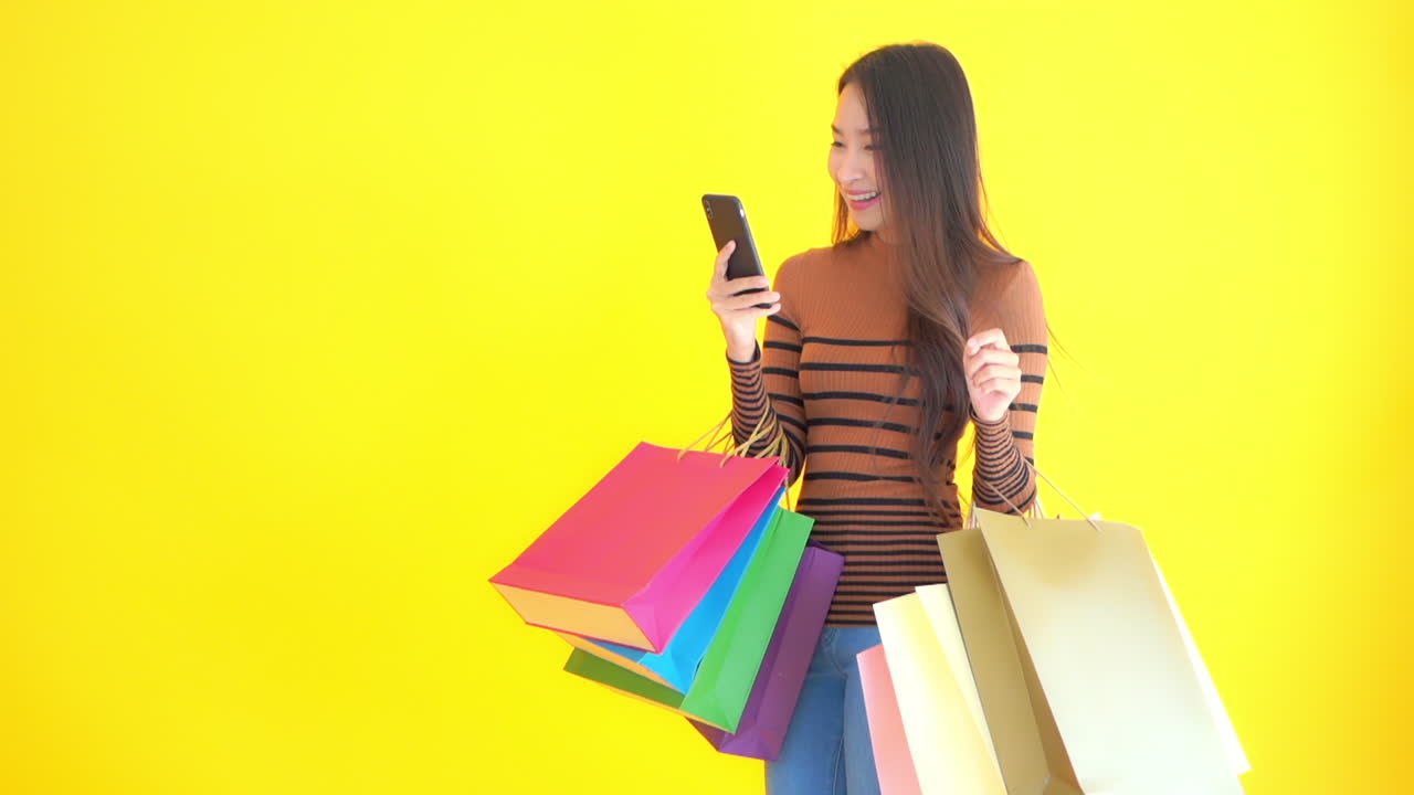 Woman with shopping bags holding smartphone in front of yellow background, copy space