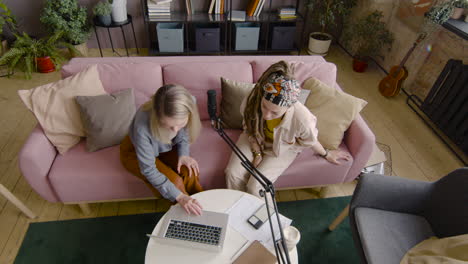 vista superior de dos mujeres grabando un podcast hablando con un micrófono mientras se sientan en un sofá frente a la mesa con una computadora portátil y documentos 1