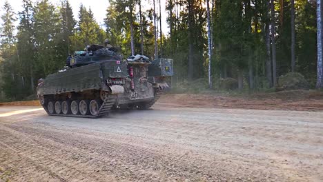 gorgeous footage of a british warrior fv510 armoured personnel carrier tank driving on a gravel road in a rural countryside sunny forest with a dust cloud behind it and soldiers with weapons on top.