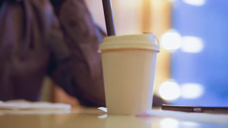 takeaway coffee cup with straw placed on table indoors beside smartphone and tissue, warm blurred lights in background with blue evening ambiance, relaxed atmosphere suggesting modern caf or casual meeting space
