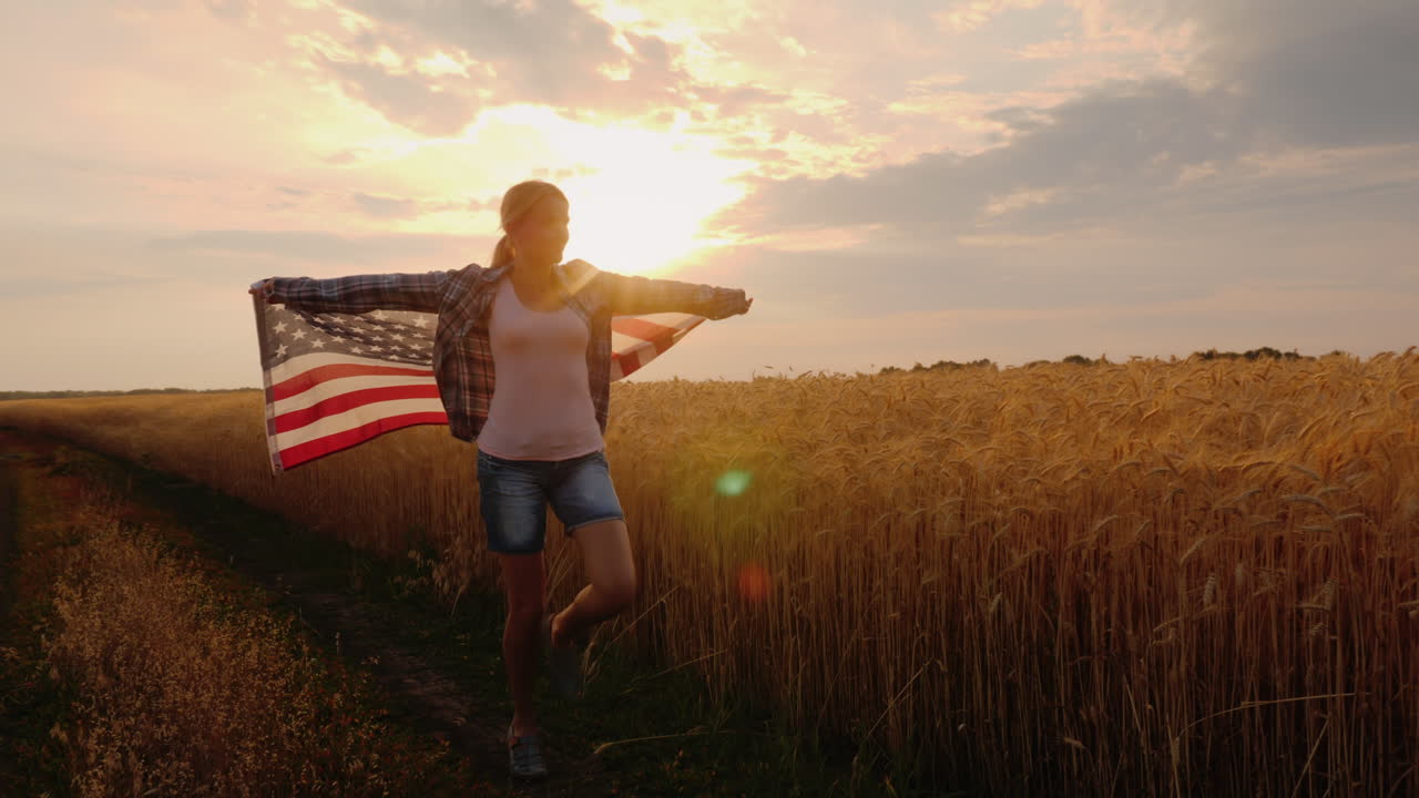 una mujer con una bandera de estados unidos atraviesa un campo de trigo bajo los rayos del sol al atardecer