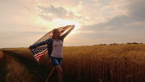 una mujer con una bandera de estados unidos atraviesa un campo de trigo bajo los rayos del sol al atardecer