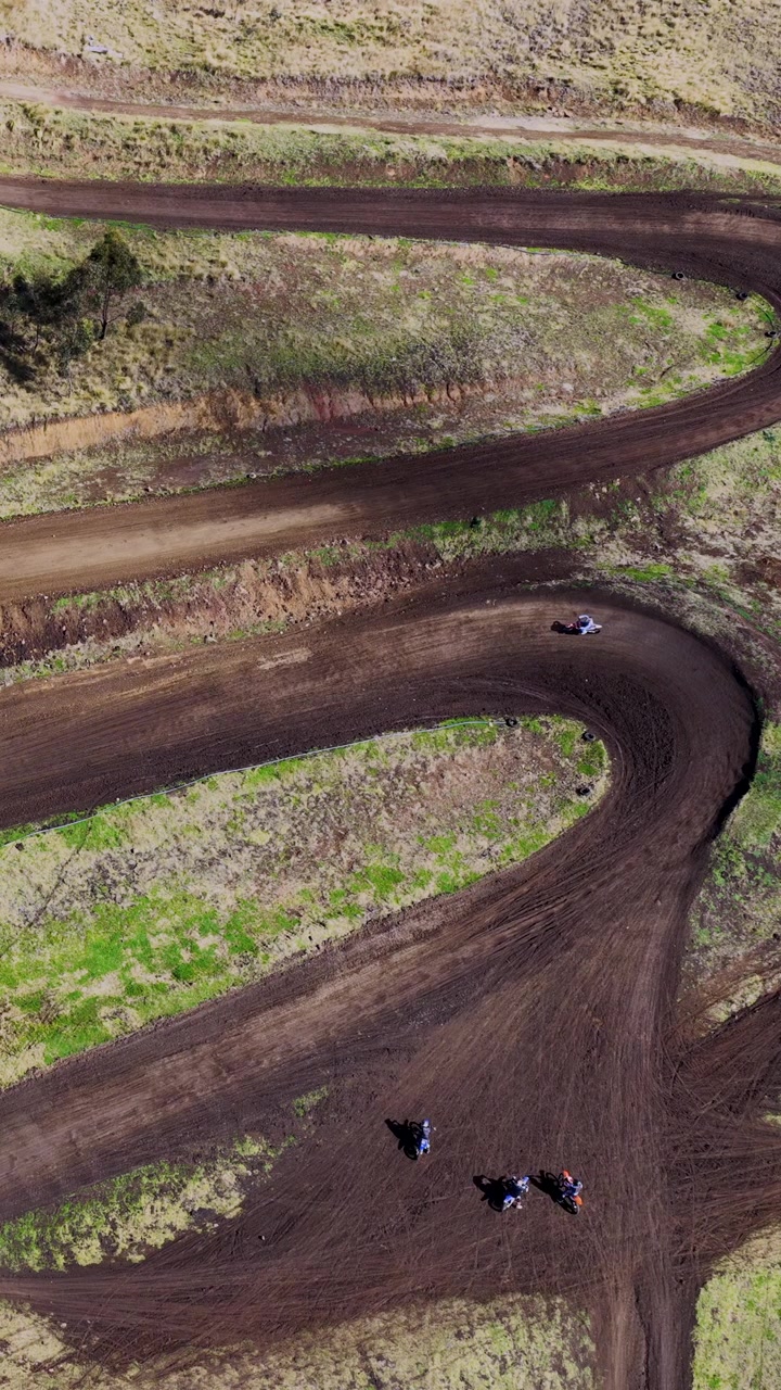 Diverse moto da cross sfrecciano su una pista da motocross fangosa e tortuosa in una boscaglia aperta, riprese dall'alto in piena luce naturale da un drone.