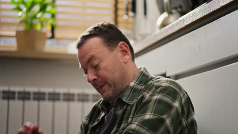 A-middle-aged-brunette-man-with-stubble-in-a-green-checkered-shirt-sighs-and-thoughtfully-puts-his-hand-on-his-head-while-solving-complex-problems-in-his-life-while-sitting-on-the-floor-in-the-kitchen